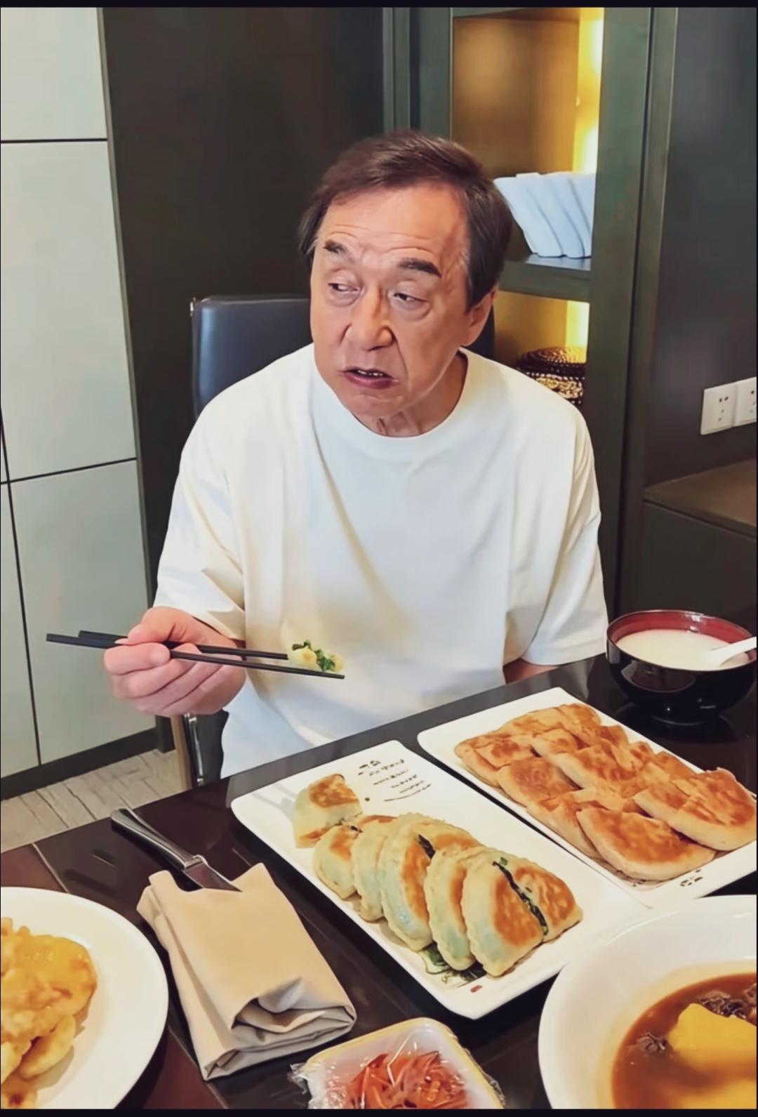 餐桌前成龙一身简单白T恤，老了有点认不出来了。他手拿筷子吃着家常菜。桌上摆满韭菜