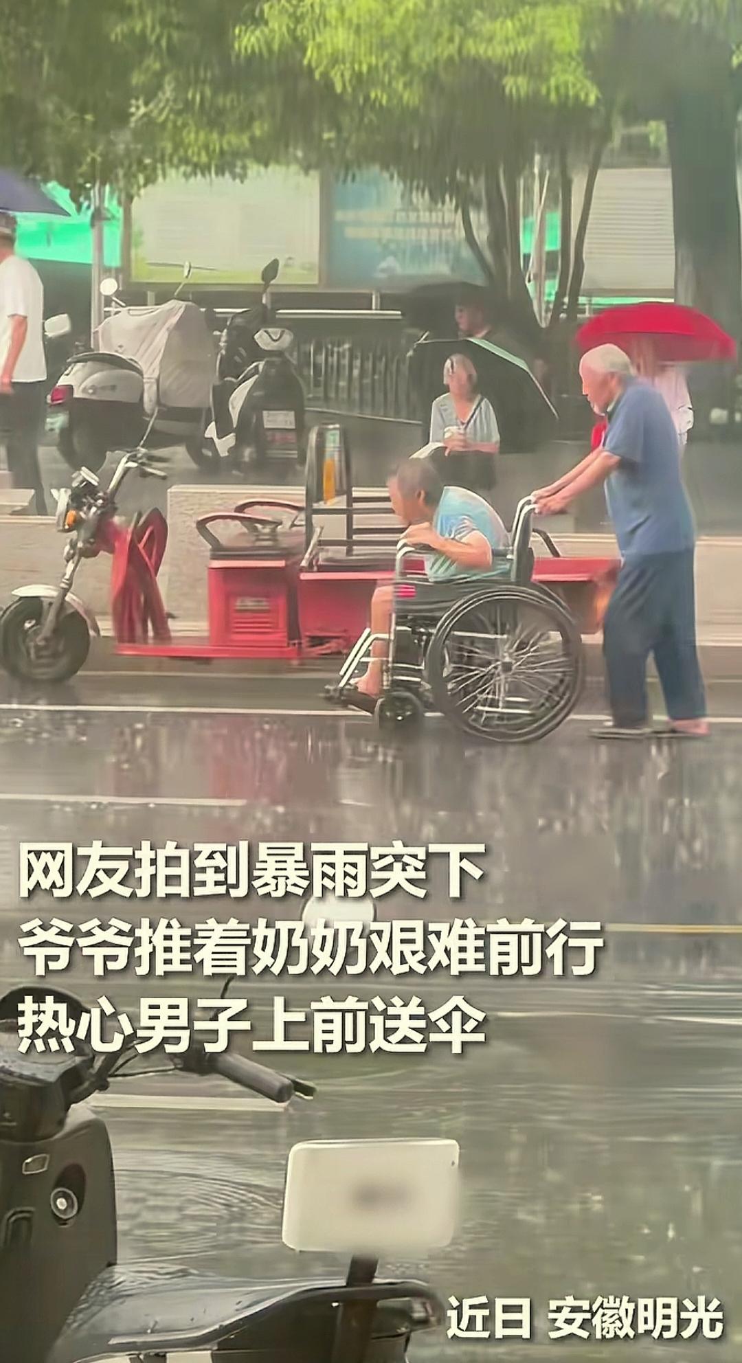 安徽明光的街头，一位白发老人用轮椅推着老伴，在大雨中艰难前行。
一位热心小伙子在