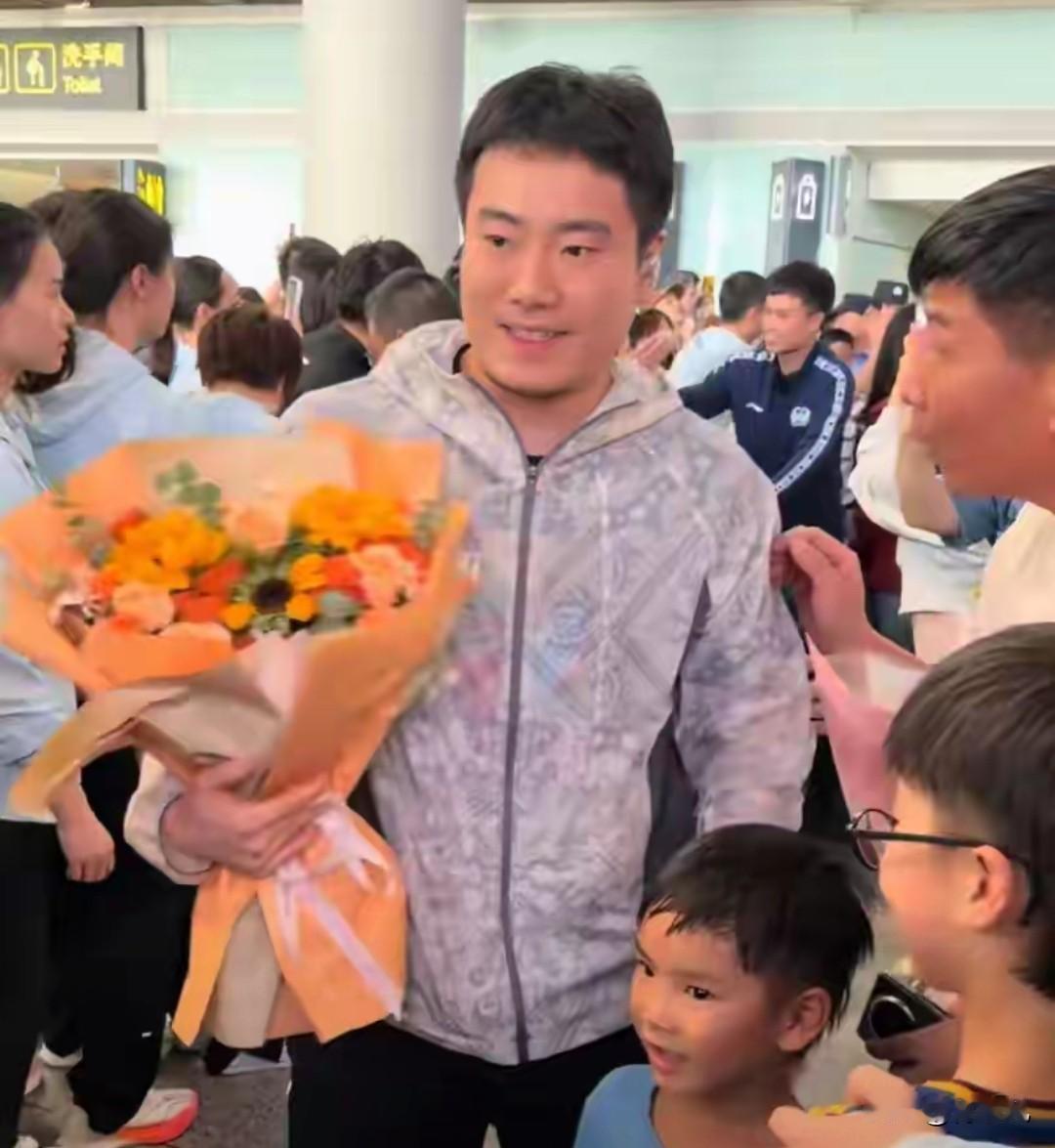 国乒载誉归来，在机场大厅，场面热烈而有序，梁靖崑的妻子和儿子为他送上鲜花，此刻，