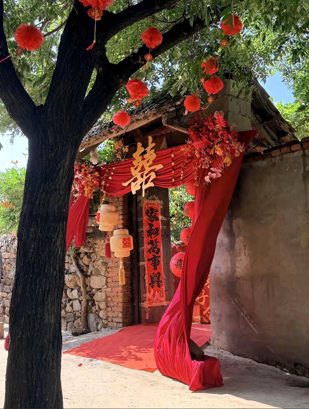 粗壮的老树枝桠间挂满红灯笼，红色布幔围成拱门，大大的金色喜字悬在门头。
石墙、旧