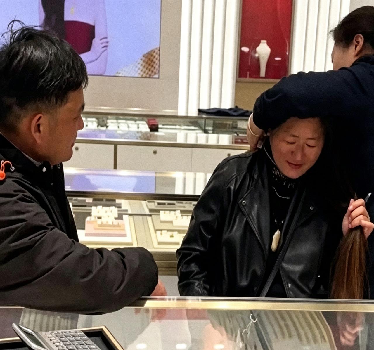 大衣哥陪大衣嫂逛金店，店员正帮忙试戴项链。大衣哥就坐对面静静瞅着，手搭在柜台上。
