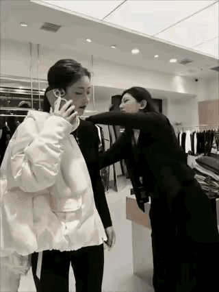 女孩在女店员的帮助下试穿衣服对着电话那头说，喂 你给我转1000块钱，我买个貂穿