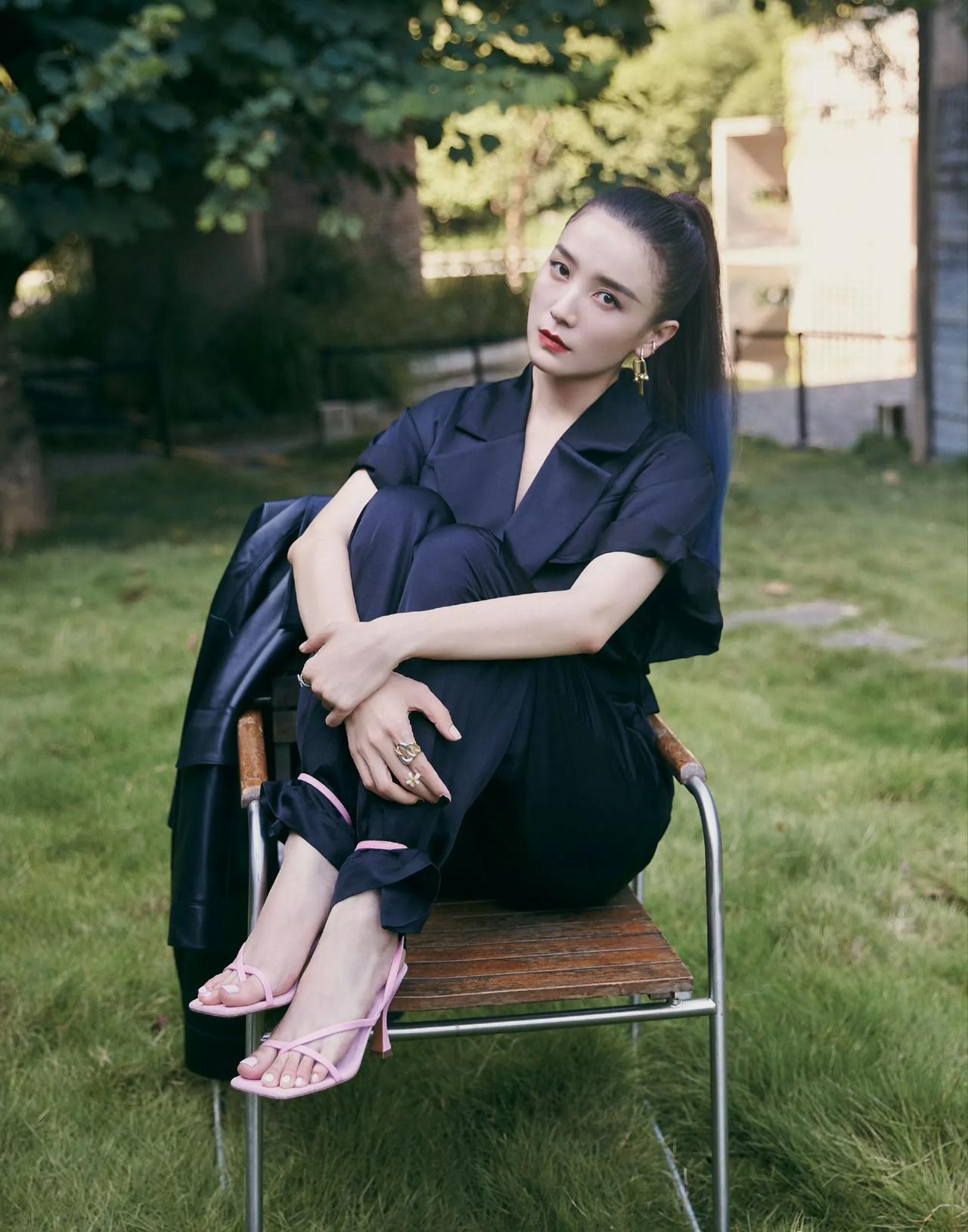绿色的草坪中，她坐在一把简单的木椅上。一身黑色的休闲衬衣套装。脸上画着精致的妆容