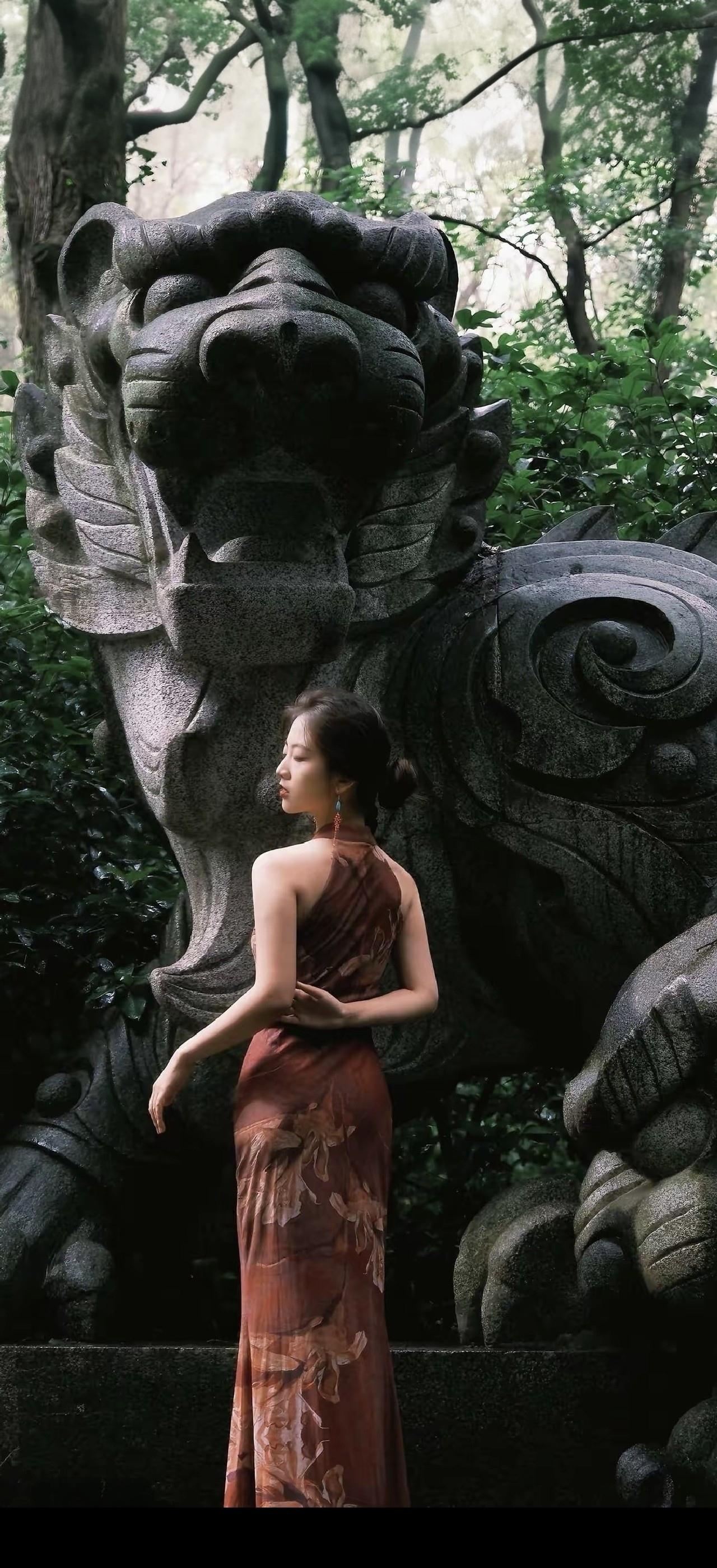 一个穿旗袍的姑娘站在石狮子旁。