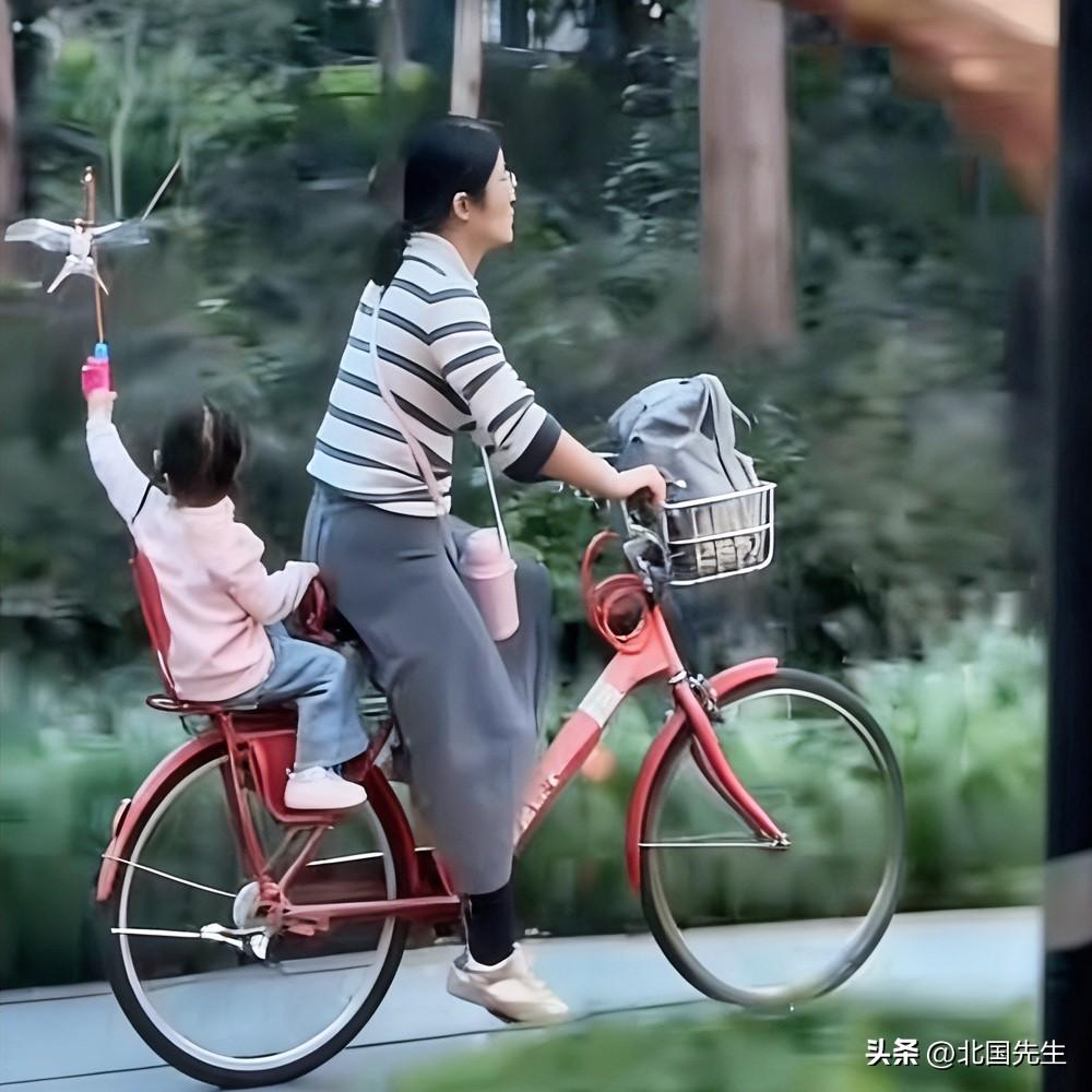 妈妈骑着标志性的“小红车”，
稳稳载着女儿穿行在春风里，

后座的小女孩高高举起