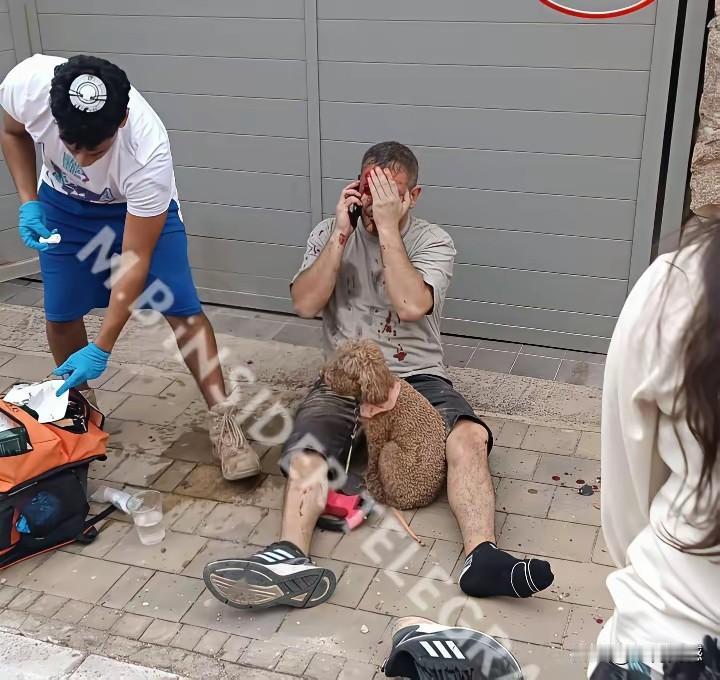 这个以色列男子在街头遭遇袭击，头部和身体都有血迹，正坐在地上接受急救，一边还在打
