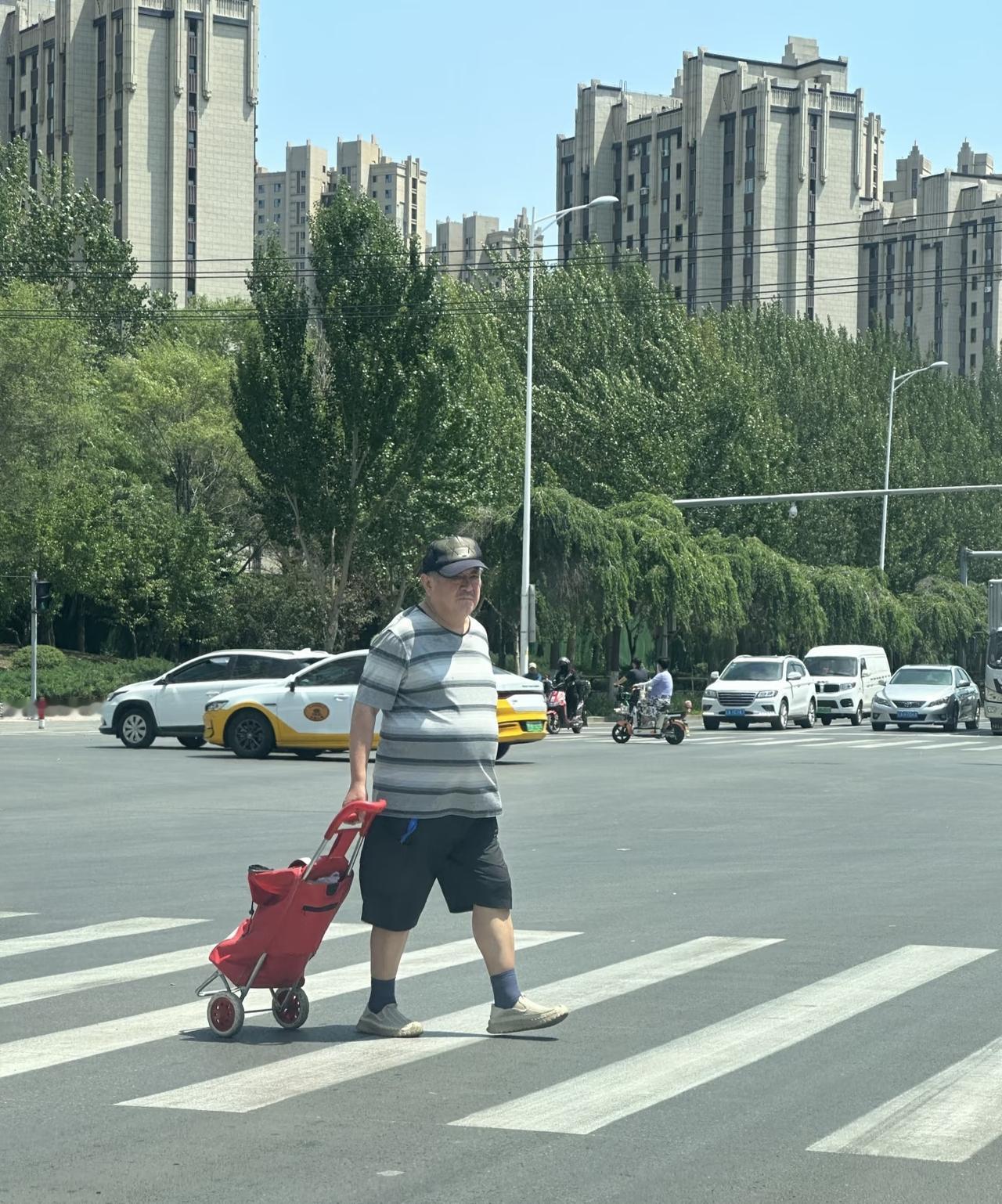 赵本山独自一人过马路，
拉着一个红色的折叠购物车，
穿着条纹T恤，
大短裤，
肚