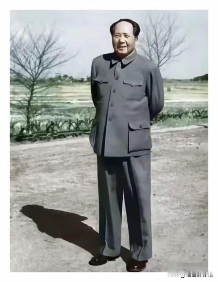 毛主席身着中山装，双手背于身后站立在郊外田野旁，神情从容舒展，面带笑容，背景是开