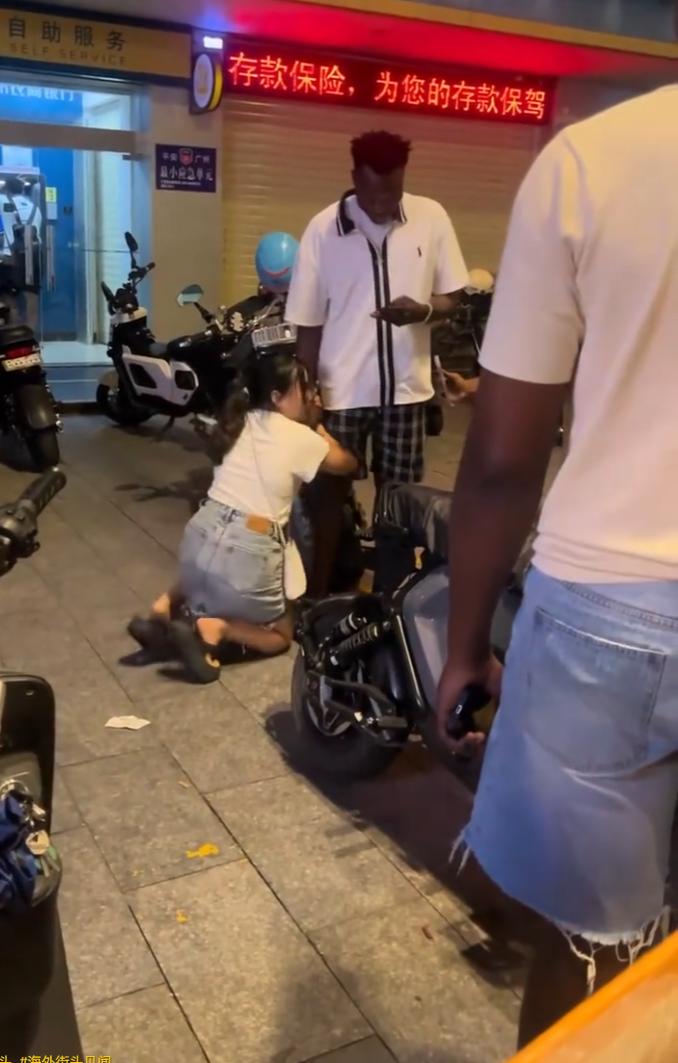 一名女子双膝跪在水泥地面，双手死死拽住黑人男子的手腕不肯松开，男人低着头盯着手机