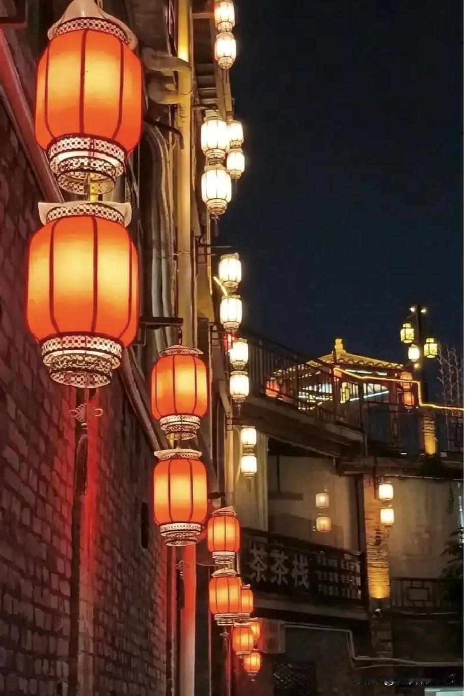 今年冬天的不同红灯笼串起夜色，暖光浸红砖墙，远处檐角也裹着光晕。巷弄里茶栈的招牌