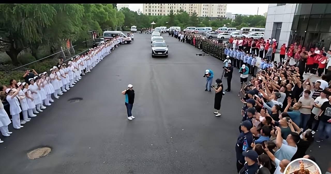 医院主干道两侧，医护、志愿者整齐分列两排肃立，道路中间留出通行通道，数十辆医疗救