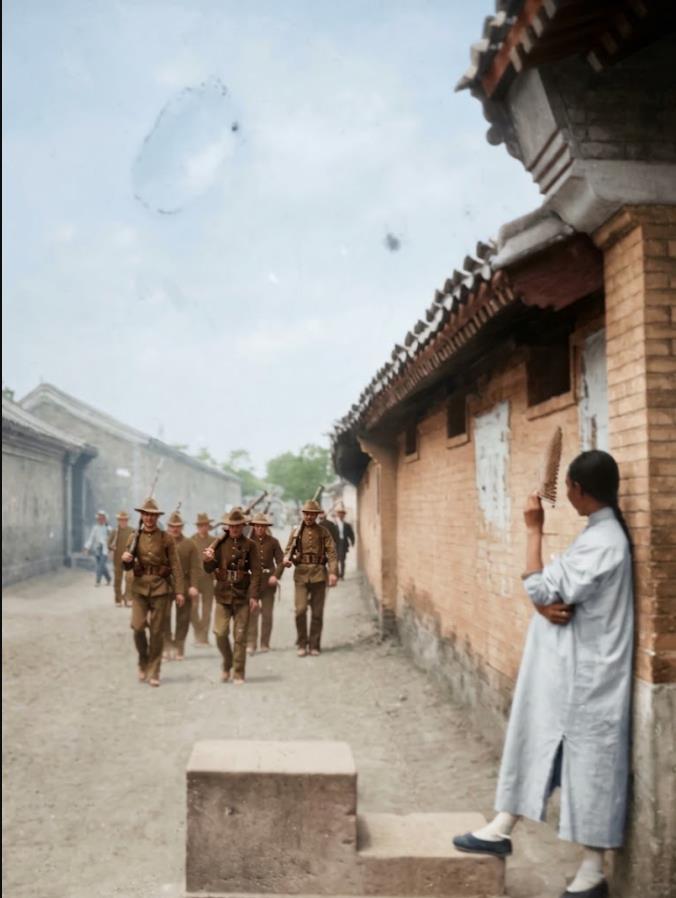 1900年，一队头戴宽檐盔、背负步枪的洋兵，列队走在北京胡同土路上。右侧砖墙门前