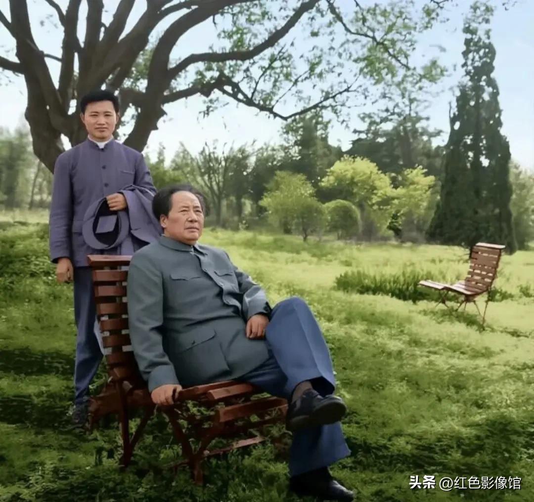 这张彩色修复照片中，毛主席静坐在园林木椅上，身旁站立工作人员，草木葱茏环境清幽，