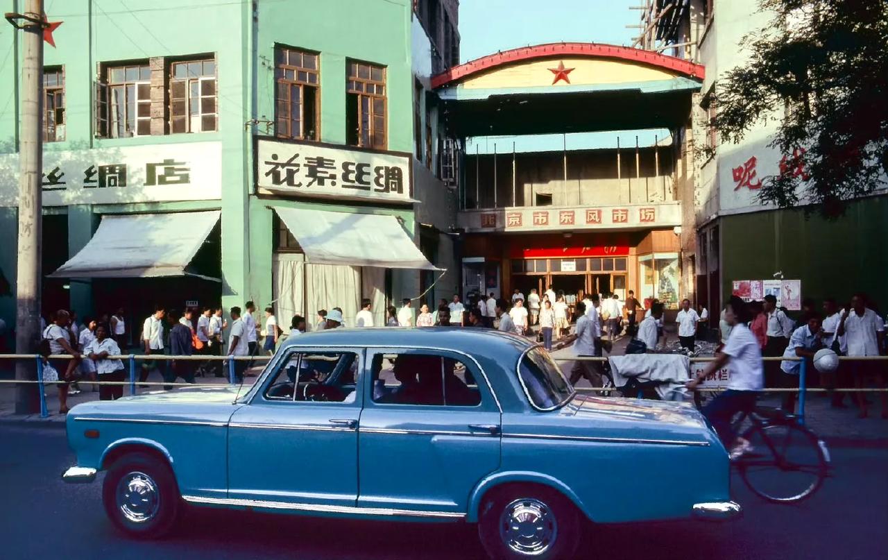 1979年夏，北京东风市门市场，其上方有红色五角星装饰，充满时代印记。周边有 “