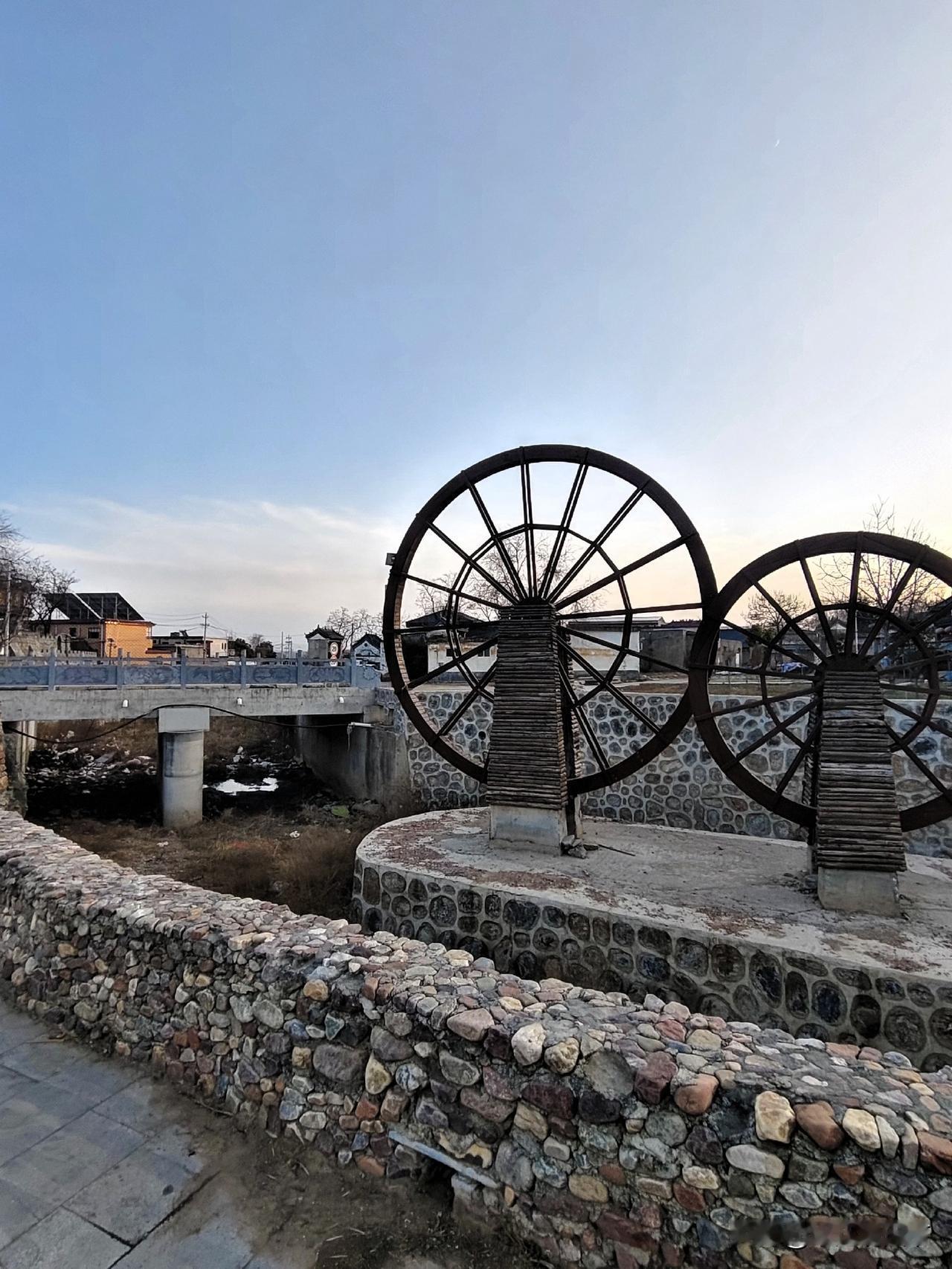 冬日的余晖斜斜洒下，落在村口那两座铁艺水车的轮辐上，勾勒出粗粝又温柔的轮廓。石砌