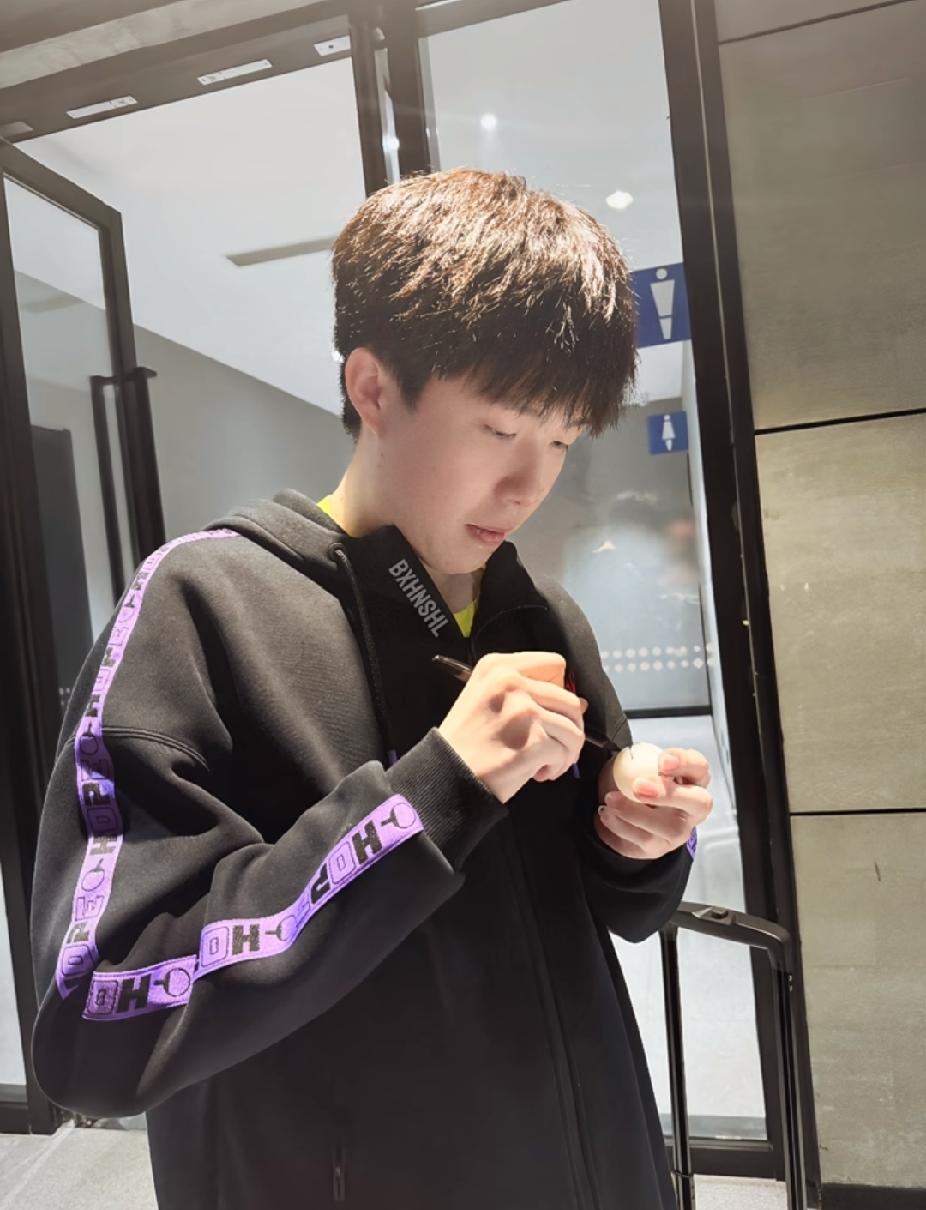 这张照片拍得真好，低头认真给乒乓球签名的样子，专注又温柔。
 
黑色连帽衫配紫色