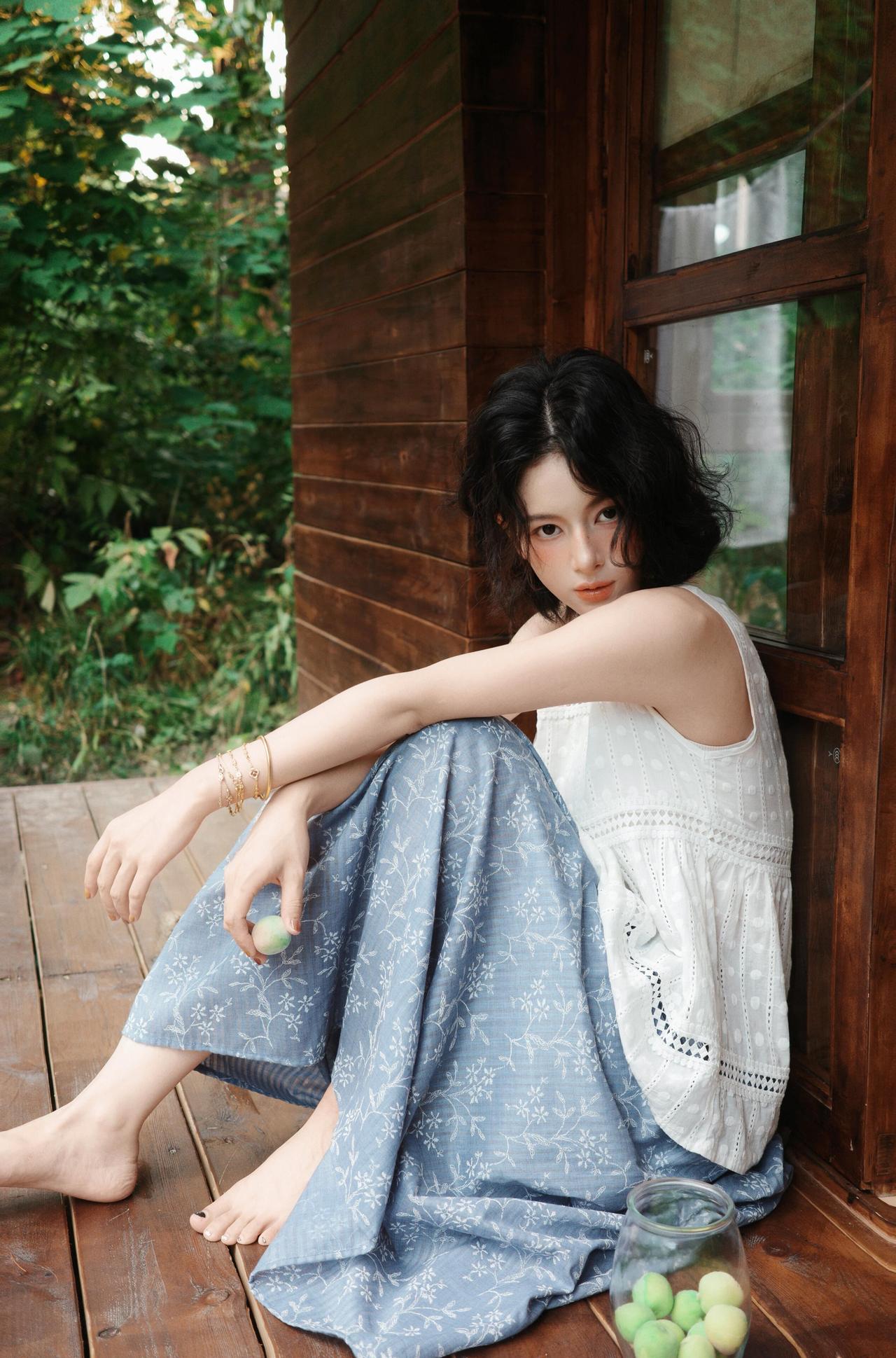 这张照片的户外场景把夏日气息展现得很鲜活。女子坐在木质建筑的角落，背后是茂密的绿