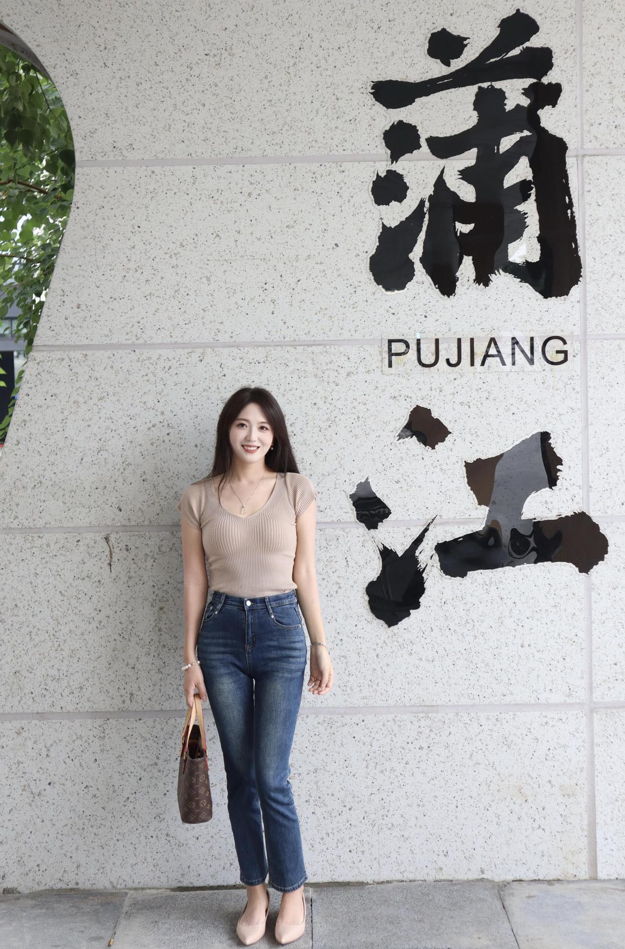 她站在“浦江”墙前，像被城市风吻过的静物画：浅棕针织衫温柔贴合肩线，牛仔蓝在高腰