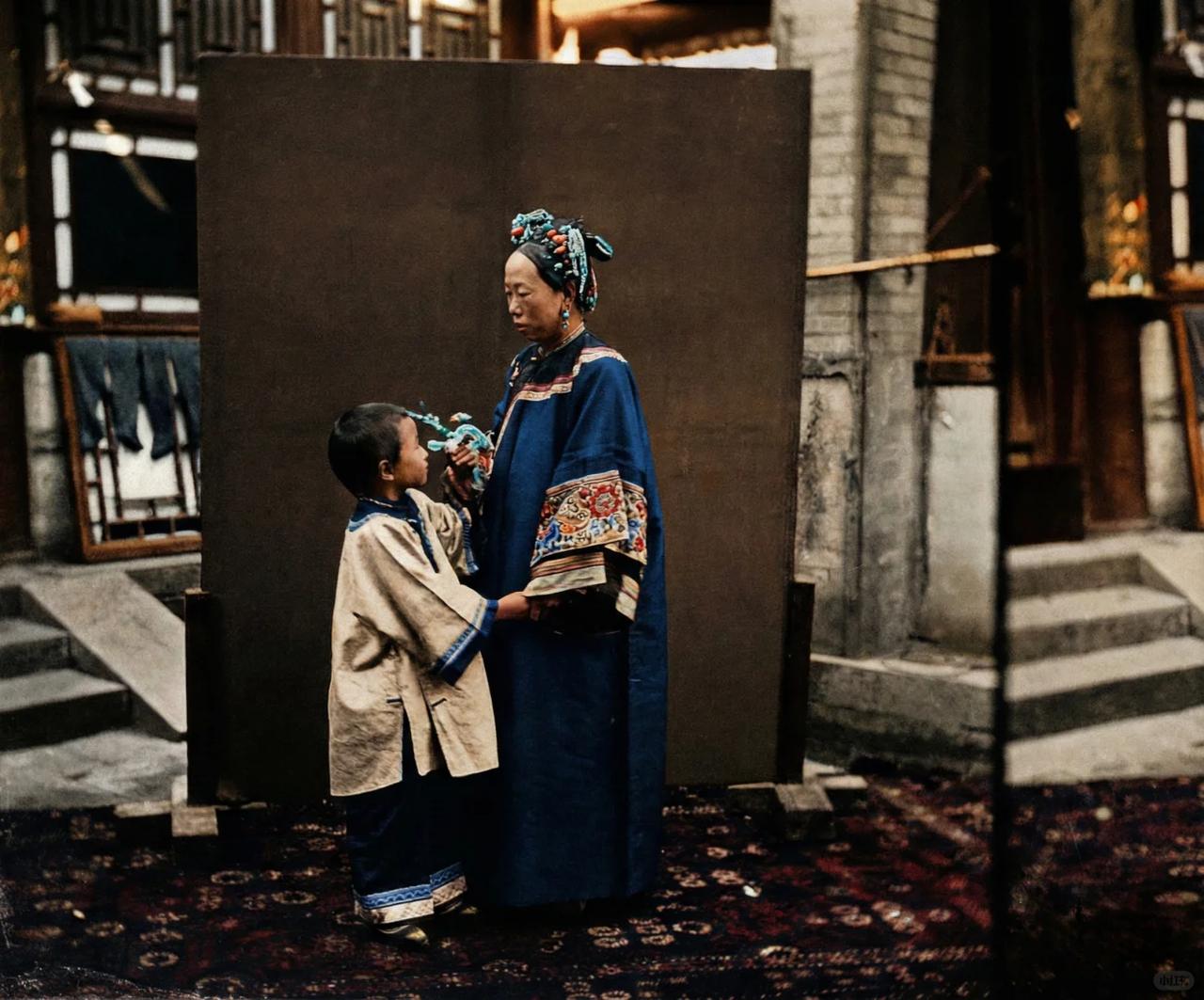 这张照片晚清简陋的街头布景里，定格了一对母子相依的温情瞬间。
画面中的母亲身着旗