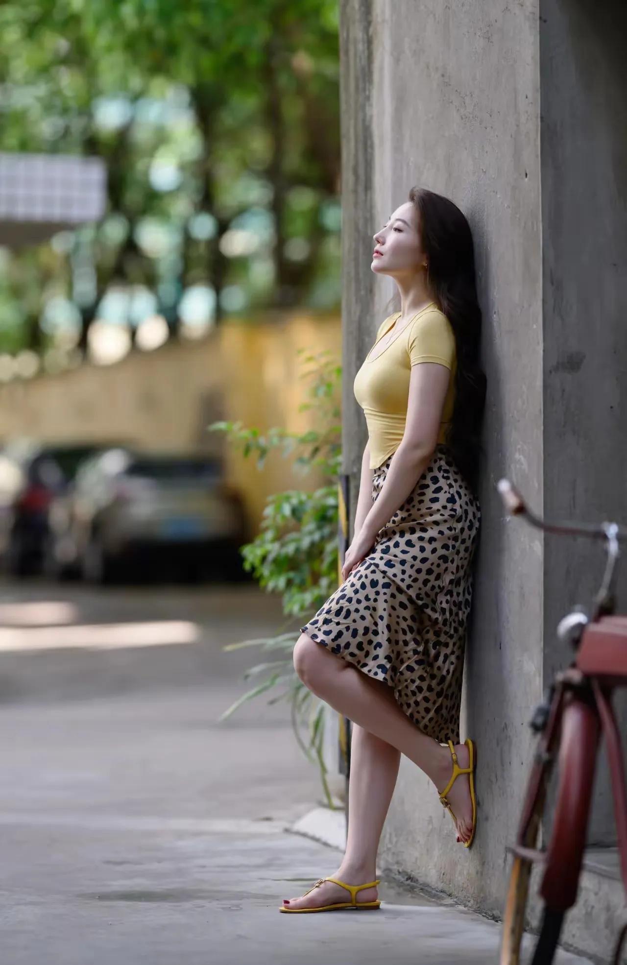 都市街头，优雅女子倚墙而立，黄衣与豹纹裙相映成趣，阳光洒落，尽显时尚与自信。微胖
