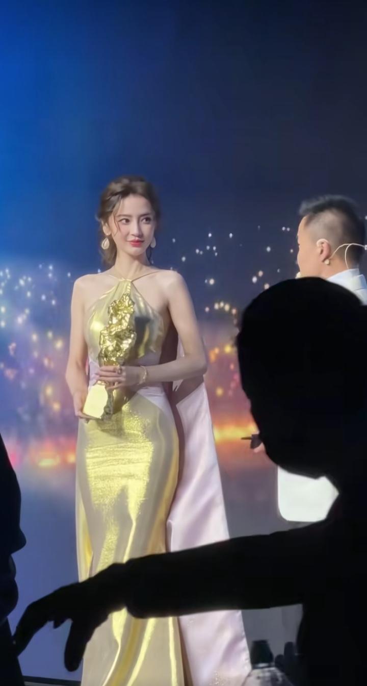 演员杨颖（Angelababy），她身着金色礼服，优雅地手持奖杯站在舞台上，灯光
