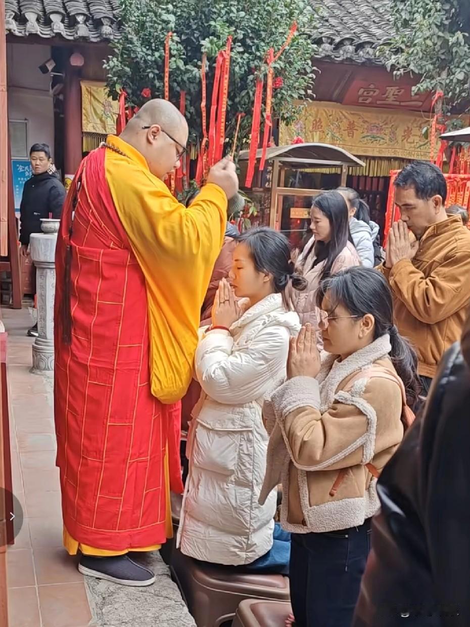 在寺庙里，一位僧人正在为信徒祈福，僧人身披红黄袈裟，手持法物静立殿前，信众俯身合