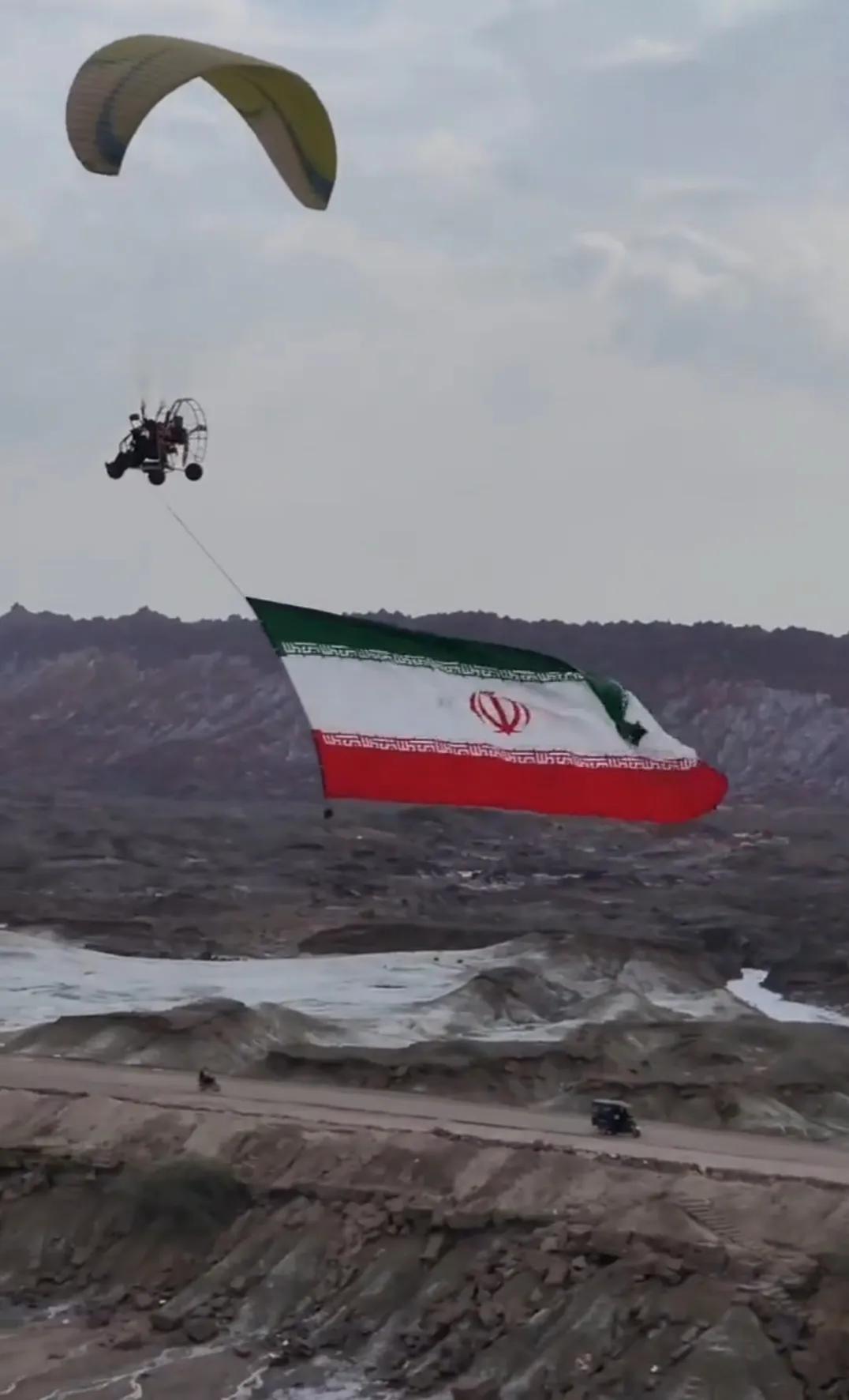 在霍尔木兹岛和霍尔木兹海峡低空动力翼伞飞行，身下的巨幅伊朗国旗迎风展开，整个画面