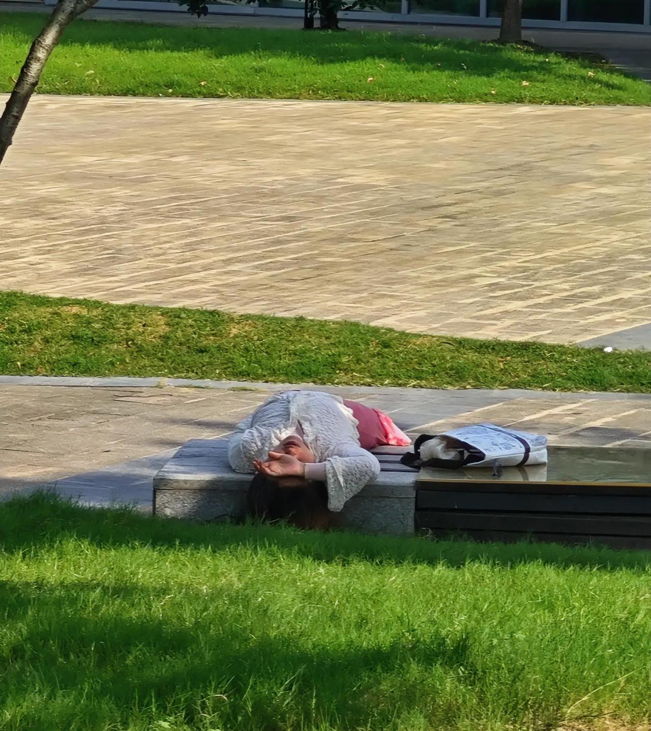 一处树荫下还算阴凉，一位身着裙装的女士，整个人平躺在公共长椅上，整张座椅全被占满