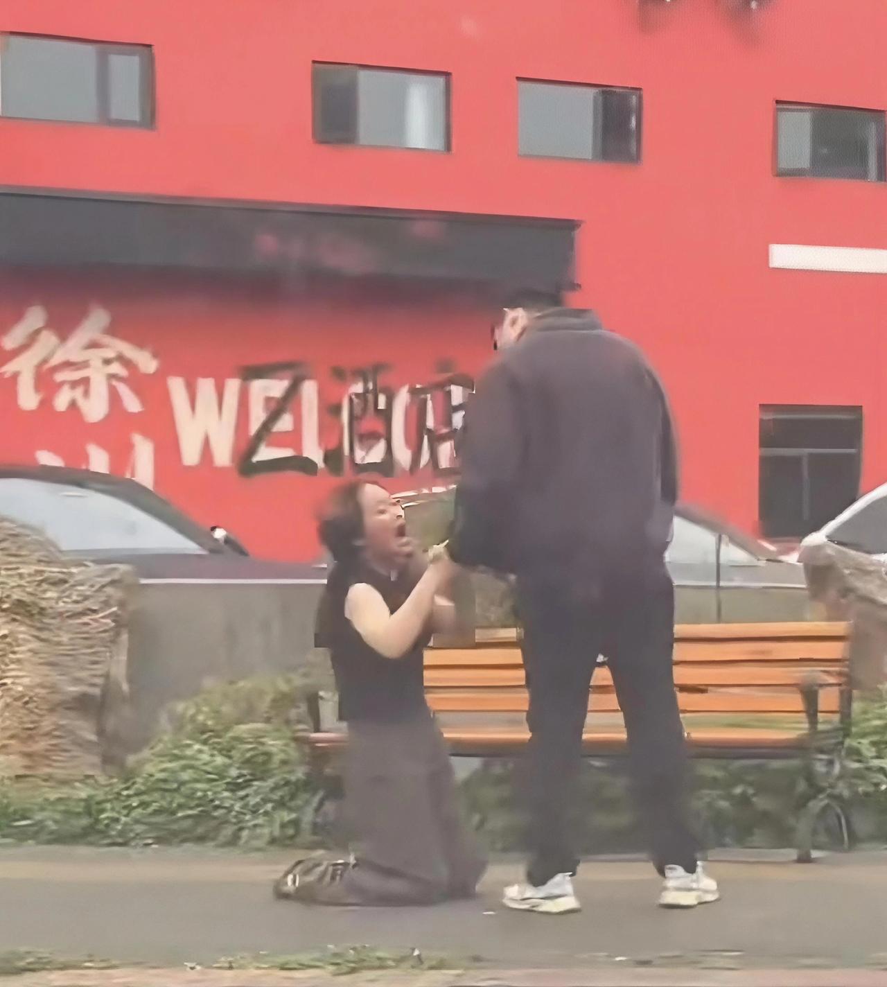 前天小雨，在徐州某地，路人拍下了这样的场景。一个女人抱着男人的双腿，不停抽打自己