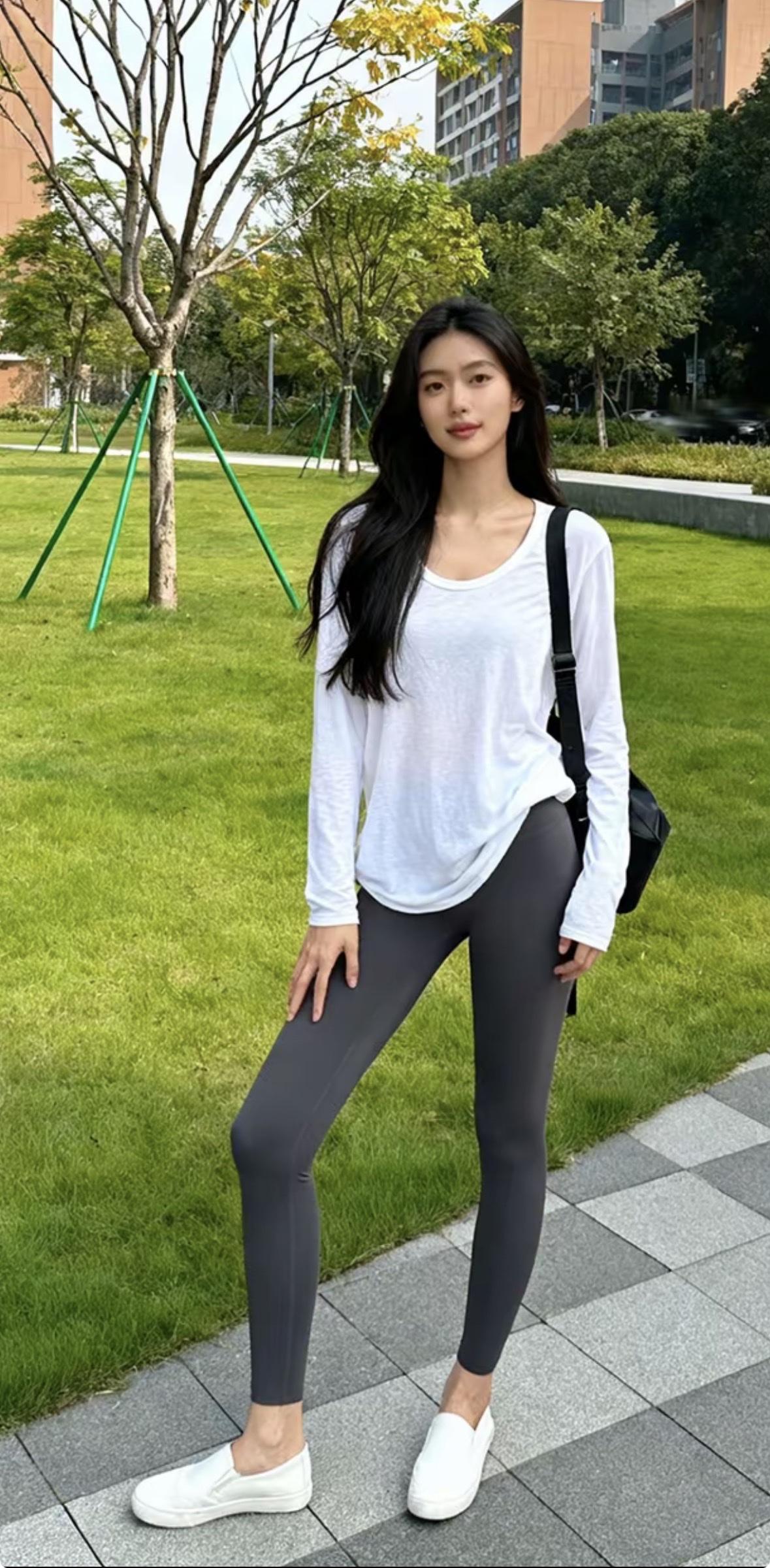 图中女子身穿白色宽松长袖上衣，搭配深灰色紧身瑜伽裤与白色平底鞋，肩背黑色单肩包。