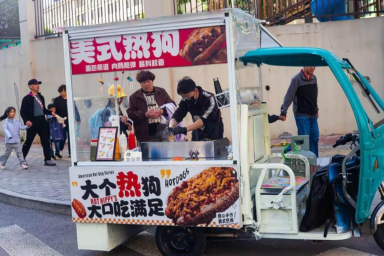 2026 年 5 月 7 日，济南宁康街，摊主在电动三轮改装的餐车上制作美式热狗