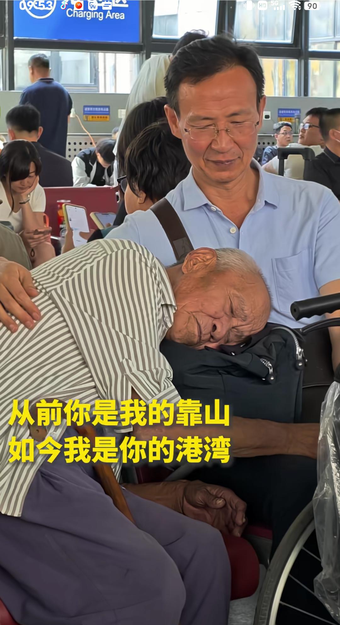 北京西站候车厅感人的一幕，儿子坐在椅子上，让年迈的父亲依偎在怀里，儿子把手耷拉着