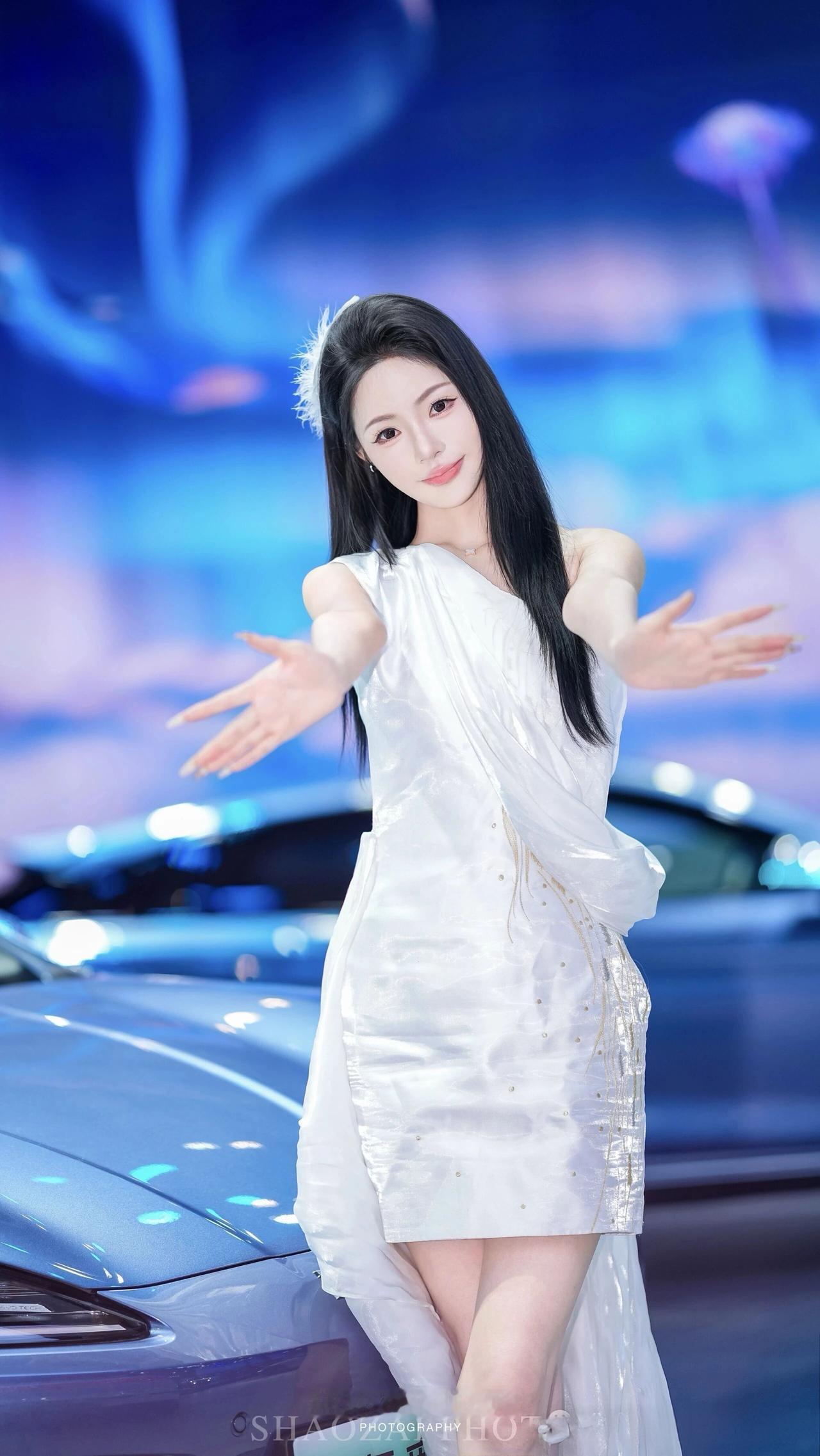 在车展的璀璨灯光里，她一袭白裙，美得自带焦点✨
穿搭与气场：单肩设计的白色长裙，