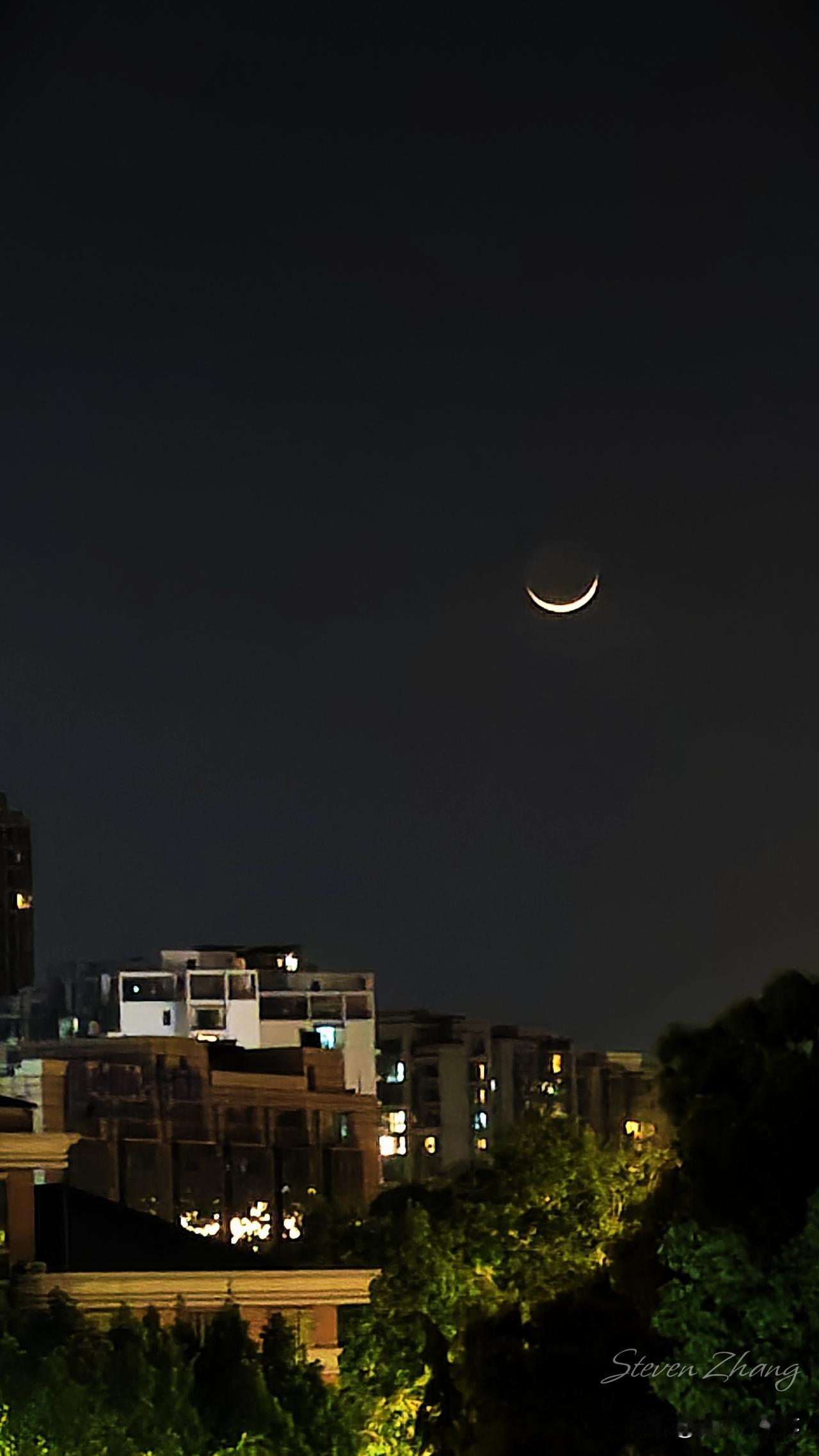 一弯低垂的月，
被温柔暮色捧在手心，
如同银钩般高挂，
枕在万家灯火之上，
或许