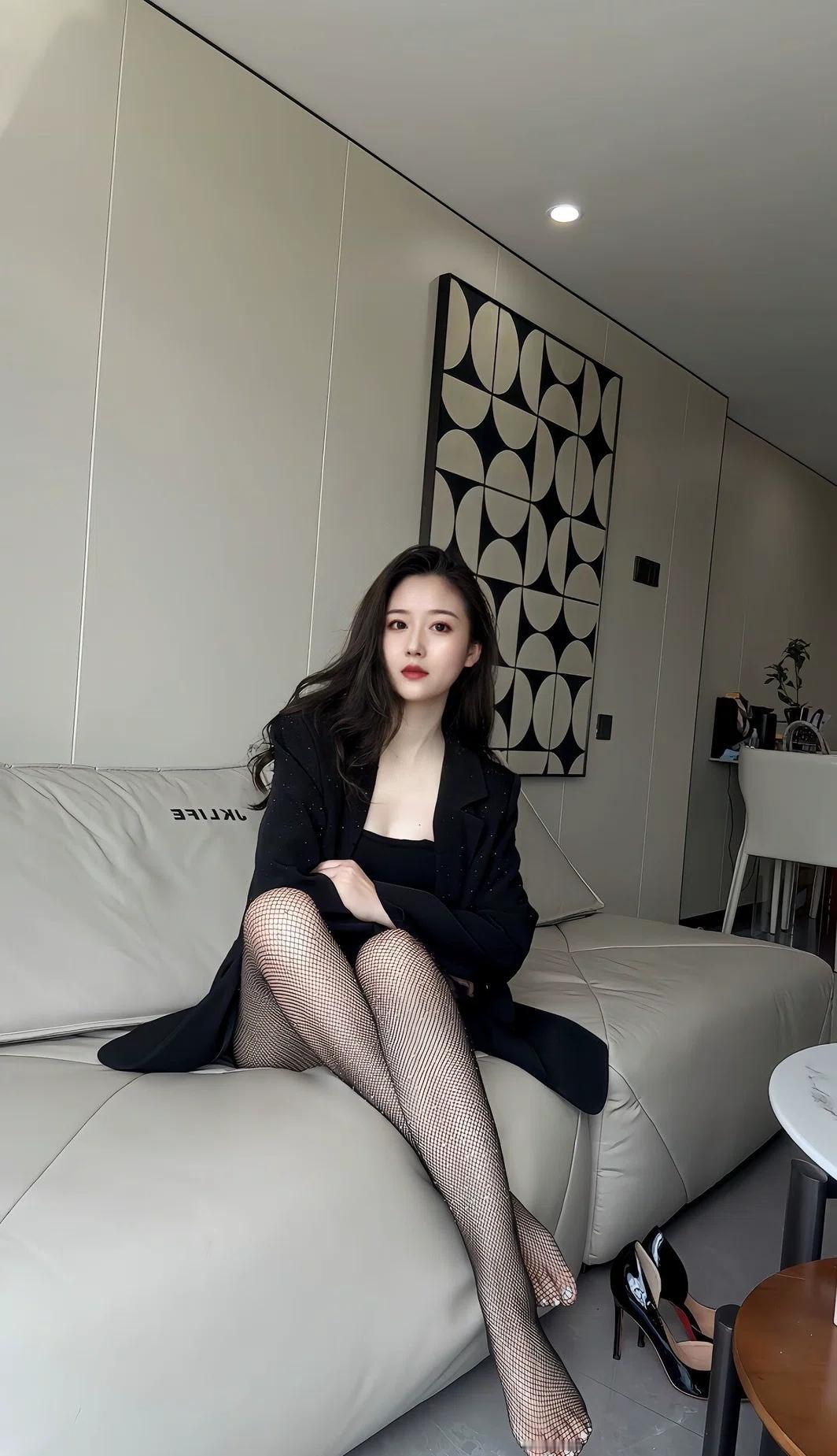 这位小姐姐坐在沙发上，穿着黑色套装和渔网袜，搭配高跟鞋，看起来非常时尚。