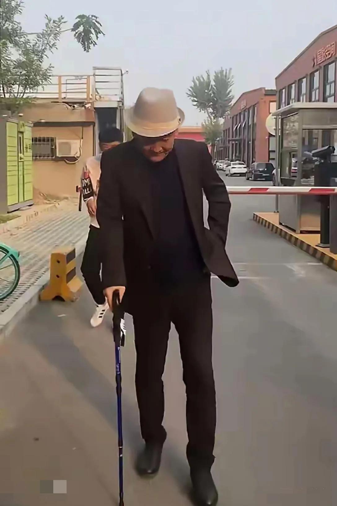 画面中的梁天体型偏瘦，穿搭讲究，气色红润，步履维艰，才66岁就拄拐杖了！！