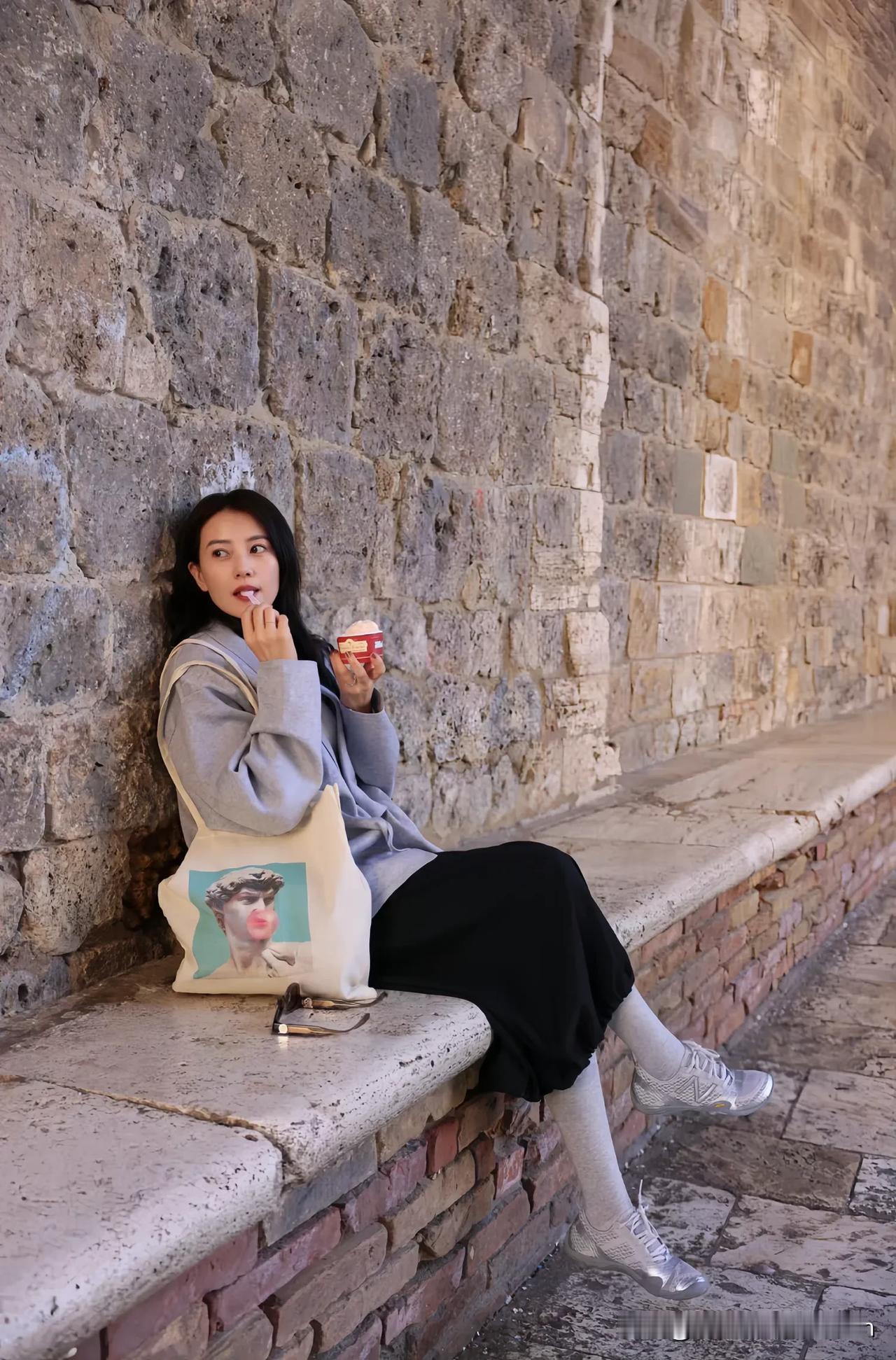 漫步在古老街巷，高圆圆倚靠着斑驳石墙，一身简约穿搭尽显随性气质。手中拿着美味甜点