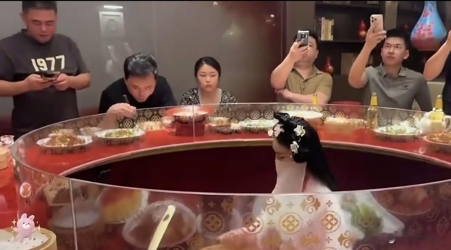 某省会大酒店包间，客人围坐边吃边喝，圆桌中心玻璃罩内，歌女弹奏古琴，供客人欣赏，