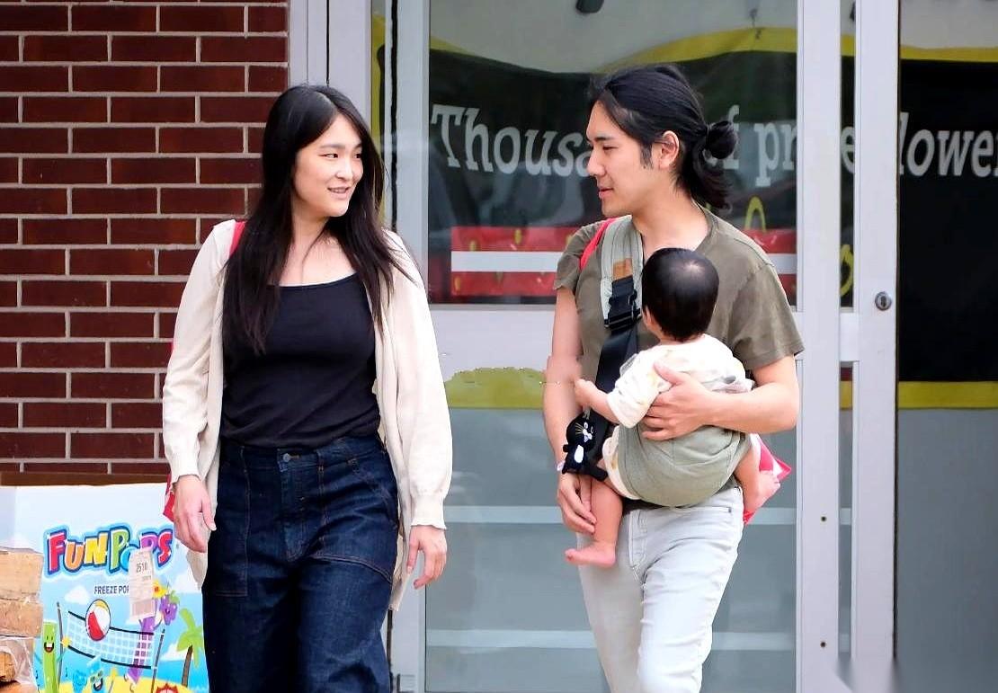 小室圭抱着娃，手里还挎着个包，那头长发甩在身后，跟旁边素颜、穿着宽松的真子公主边