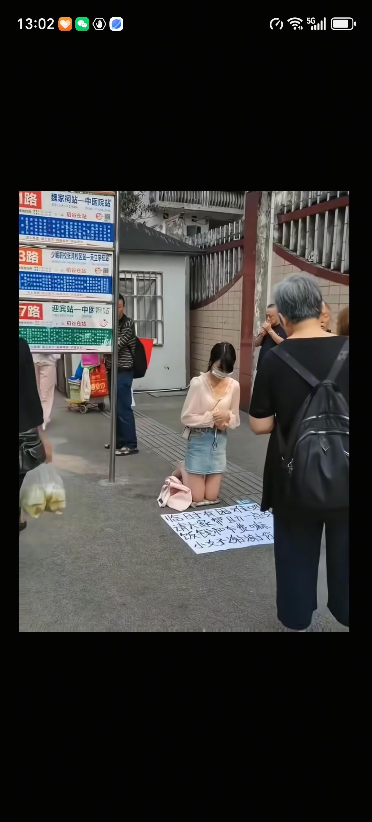 女子在街头很显眼的站牌底下跪着，光着腿，膝盖底下垫了垫子，面前一张大白纸上表明自