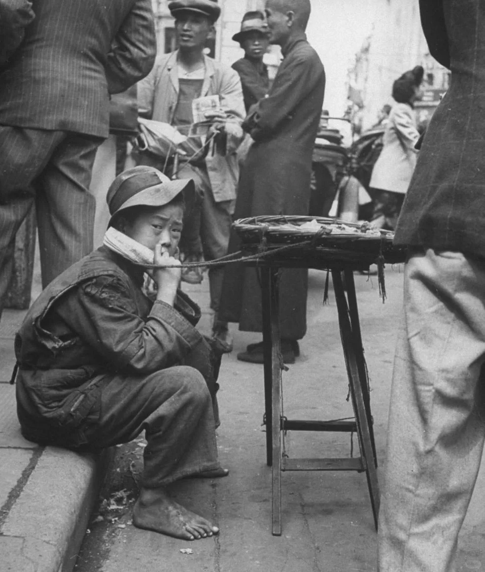 1945年11月，上海。一个小男孩坐在路边卖糖果，他身后一个时髦的青年...
