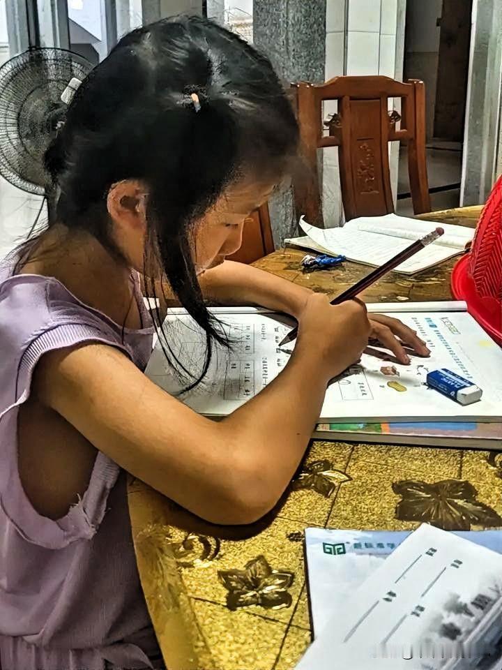 傍晚，家里的灯光柔和。小女孩穿着淡紫色的衣服，坐在铺着金色花纹桌布的桌前认真写作