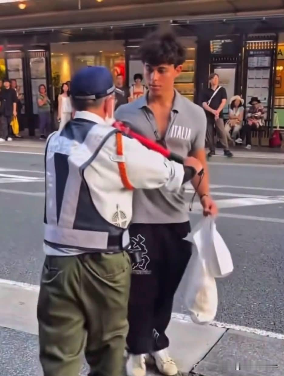 一名拎着购物袋的外籍男性游客，在日本街头想要横穿马路。现场交通疏导员看到后立刻抬