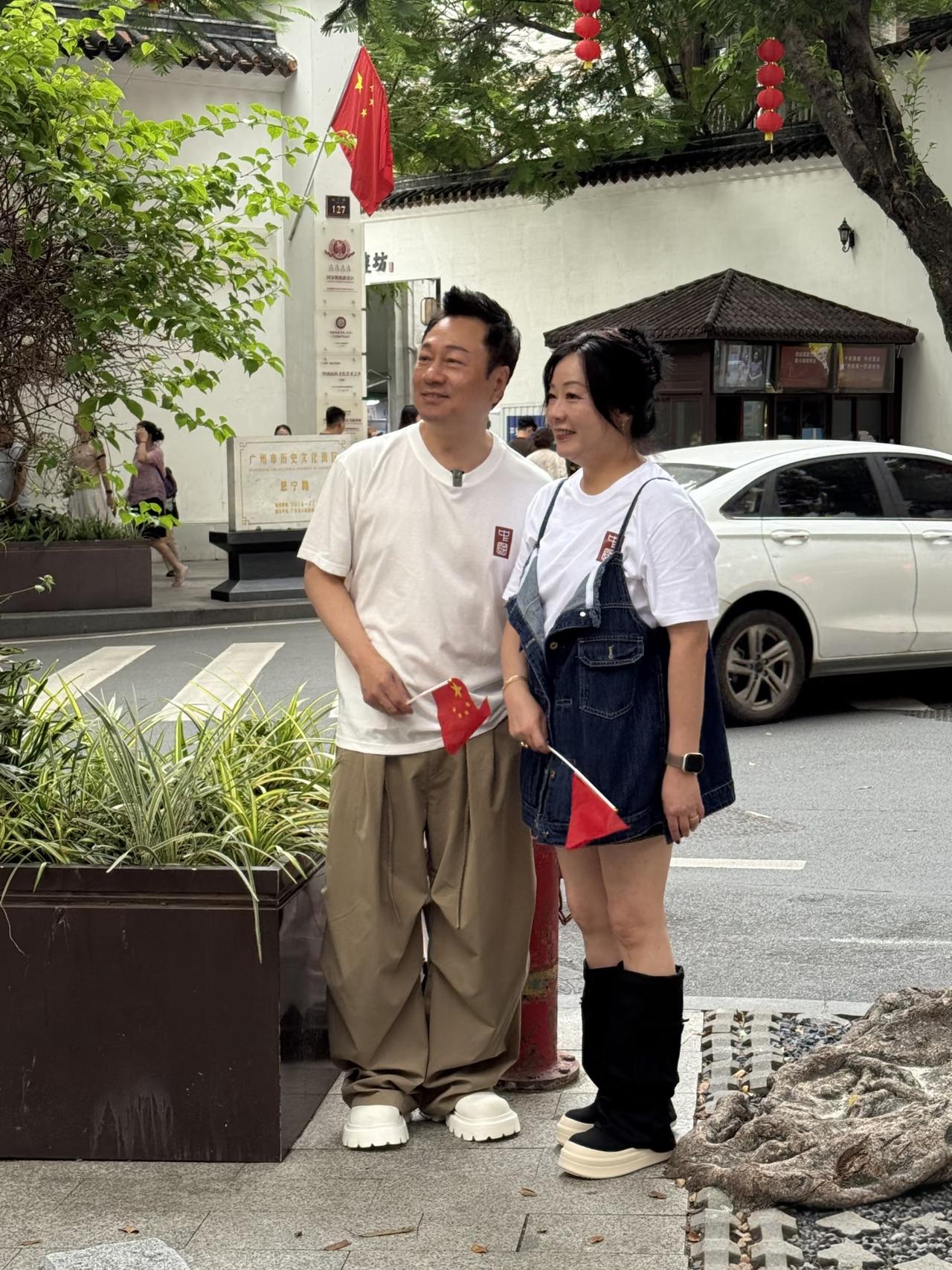 黎耀祥和老婆梁耀莲现身广州永庆坊旅游，两个人站在路边，手里拿着小五星红旗，庆祝即