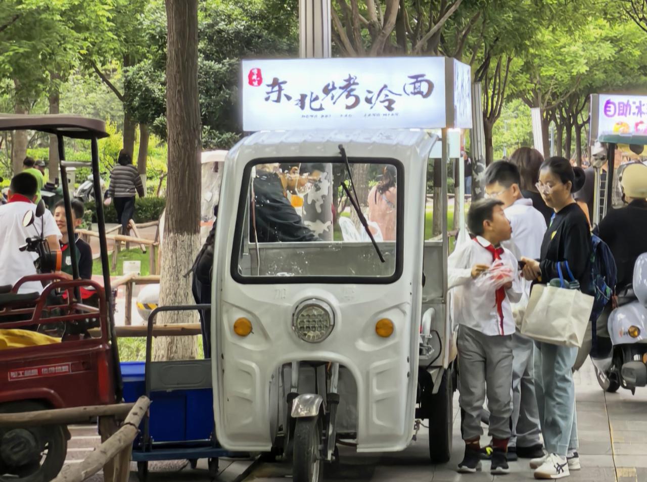 梧桐树下的白色小车，藏着小区妈妈默许的“放学补给站”——东北烤冷面一热，酱香混着