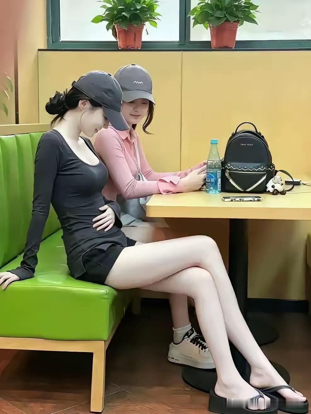 绿皮软包卡座上，两位戴着同色系棒球帽的女生挨坐在一起，深灰紧身衣配黑短裙的那位手