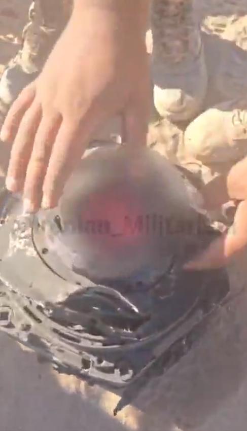 视频中几只手在沙地上摆弄着受损的金属残骸，这是一个带有彩色透镜的球形传感器，估计