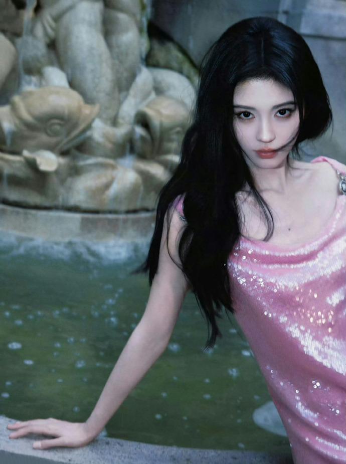 鞠婧祎
喷泉的水纹漫过石沿
像一整池被揉碎的星
她坐在冷硬的边缘
粉色亮片裙，是