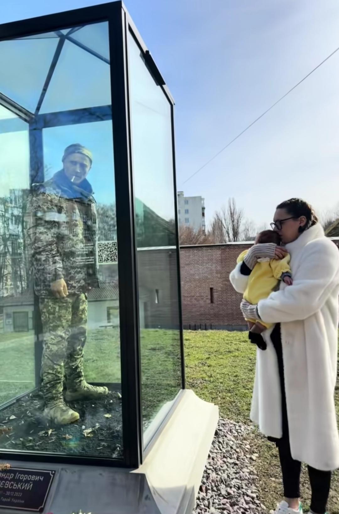 一名乌克兰妇女抱着小孩，祭拜战死的老公，小孩着父亲的雕像不知道他是谁，世间悲伤莫