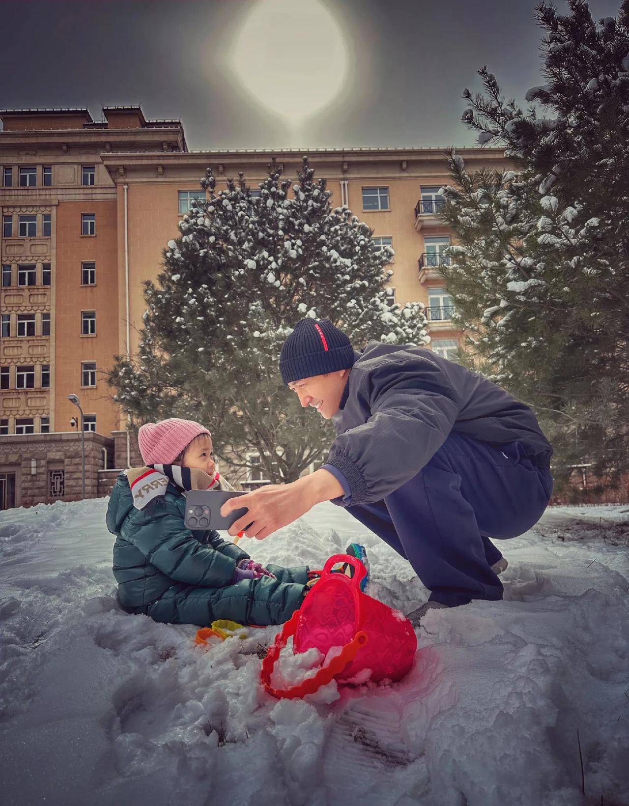 演员王阳和女儿在雪地里玩耍，享受温馨的亲子时光。女儿身穿绿色羽绒棉衣，戴着粉色帽
